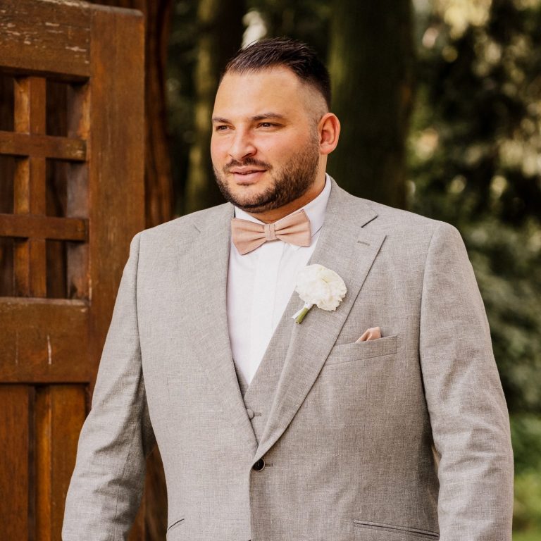Mann in einem grauen Anzug mit Fliege und Blumenboutonniere, steht vor einer Holzstruktur.